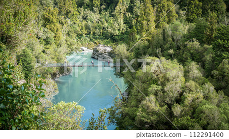 People crossing swing bridge above gorge with azure waters surrounded by dense forest, New Zealand People crossing swing bridge above gorge with azure waters surrounded by dense forest, New Zealand 112291400