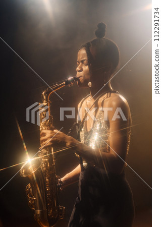 Vertical portrait of Black young woman performing jazz music on stage and playing saxophone retro style with gold accent light Vertical portrait of Black young woman performing jazz music on stage and playing saxophone retro style with gold accent light 112291734