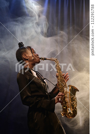 Vertical portrait of talented Black woman playing saxophone on stage during performance in jazz music club with smoke effect 112291788