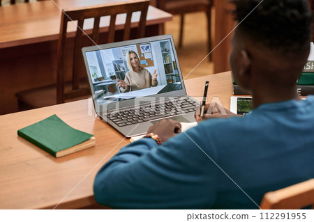 Back view of young Black man watching video lecture in school library and using laptop with teacher speaking on screen Back view of young Black man watching video lecture in school library and using laptop with teacher speaking on screen 112291955
