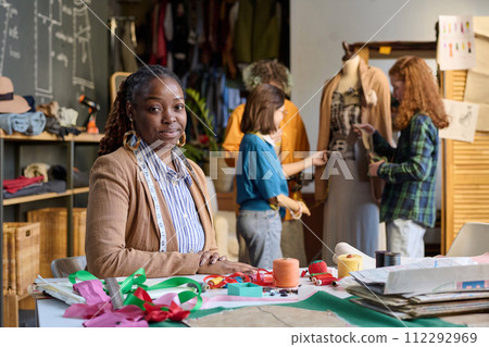 Portrait of African American adult woman sitting at desk in atelier workshop and looking at camera with girls in background copy space 112292969
