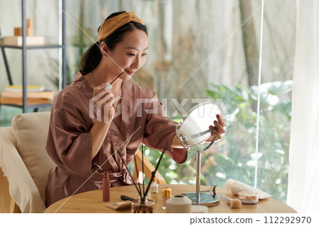 Smiling Chinese young woman applying makeup when getting ready for date or event Smiling Chinese young woman applying makeup when getting ready for date or event 112292970