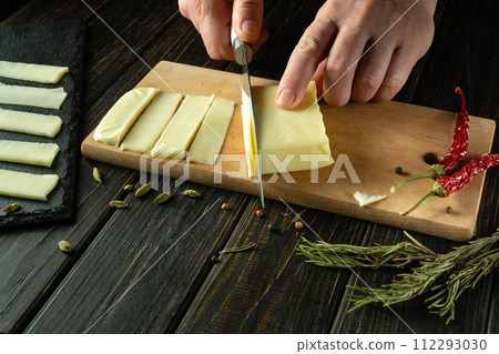 The cook uses a knife to cut Mozzarella cheese into thin strips for table setting. Low key concept of serving a dairy dish for lunch The cook uses a knife to cut Mozzarella cheese into thin strips for table setting. Low key concept of serving a dairy dish for lunch 112293030