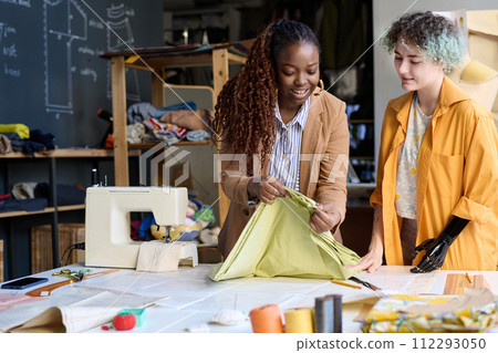 Waist up portrait of young woman with disability sewing clothing in inclusive atelier with smiling mentor examining work copy space 112293050