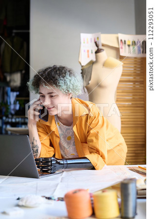 Vertical portrait of young woman with arm prosthesis speaking by phone and using laptop managing atelier business 112293051