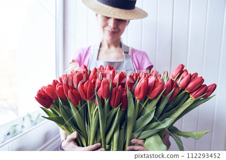 Woman holding a big bouquet of red tulips with a copy space. Gardener wearing a hat and holding beautiful flowers in the greenhouse. Woman holding a big bouquet of red tulips with a copy space. Gardener wearing a hat and holding beautiful flowers in the greenhouse. 112293452