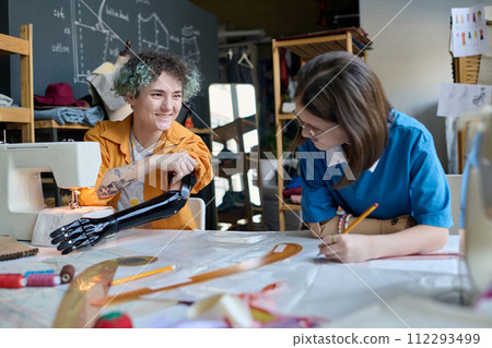 Portrait of two smiling teenage girls with prosthetics sewing clothing together in inclusive training class Portrait of two smiling teenage girls with prosthetics sewing clothing together in inclusive training class 112293499