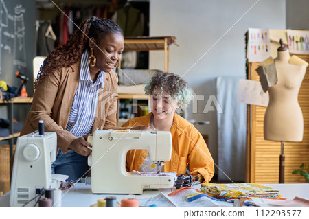 Portrait of smiling teen girl with disability using sewing machine in vocational training class with social worker helping copy space 112293577
