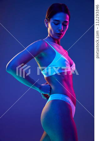 Focusing on fit and form. Young and slim woman in white lingerie posing in multicolored neon light against gradient blue studio background 112293846