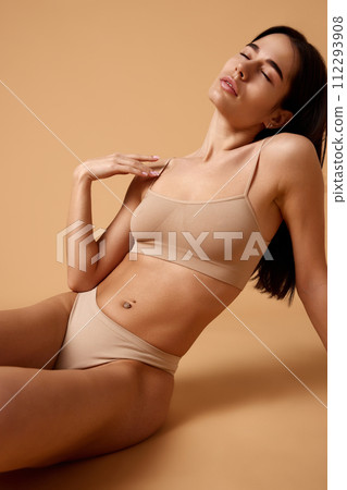 Joyful, relaxed young woman sitting in beige lingerie on floor and posing against sandy color studio background. Joyful, relaxed young woman sitting in beige lingerie on floor and posing against sandy color studio background. 112293908