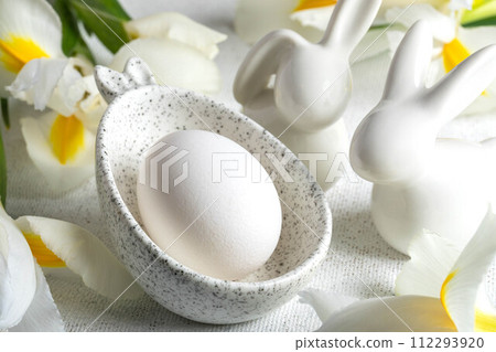 One Easter egg in marble bowl with bunny ears, small bunnies, white Iris flowers on white close up. One Easter egg in marble bowl with bunny ears, small bunnies, white Iris flowers on white close up. 112293920