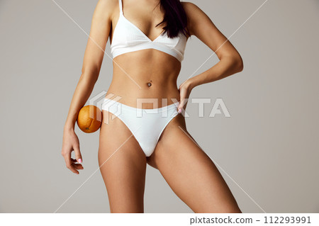 Woman's lower body in white underwear holding an orange near hip against grey studio background. 112293991
