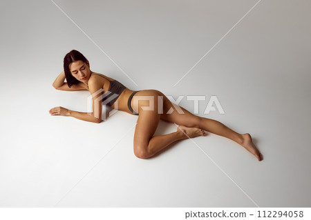 View from above of young woman posing in lingerie shows her perfect, slim body lying on floor against grey studio background. View from above of young woman posing in lingerie shows her perfect, slim body lying on floor against grey studio background. 112294058