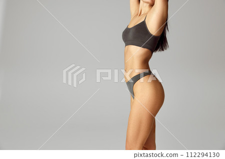 Cropped portrait of young woman with slim figure posing in lingerie raising arms against grey studio background. Cropped portrait of young woman with slim figure posing in lingerie raising arms against grey studio background. 112294130