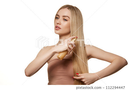 Beautiful young woman taking care after long, shiny blonde hair, applying hair spray, liquid conditioner and heat protection isolated against white studio background 112294447