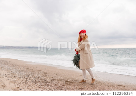 Redhead woman Christmas tree sea. Christmas portrait of a happy redhead woman walking along the beach and holding a Christmas tree in her hands. She is dressed in a light coat and a red beret. 112296354