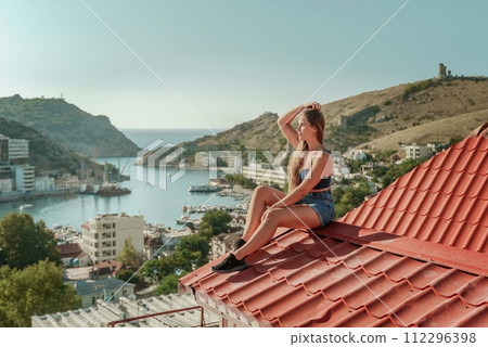 Woman sits on rooftop, enjoys town view and sea mountains. Peaceful rooftop relaxation. Below her, there is a town with several boats visible in the water. Rooftop vantage point. 112296398