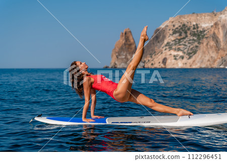 Sporty girl on a surfboard in the sea on a sunny summer day. In a red swimsuit, she sits in the splits on the sap. Summer entertainment on Stortom by the sea Sporty girl on a surfboard in the sea on a sunny summer day. In a red swimsuit, she sits in the splits on the sap. Summer entertainment on Stortom by the sea 112296451