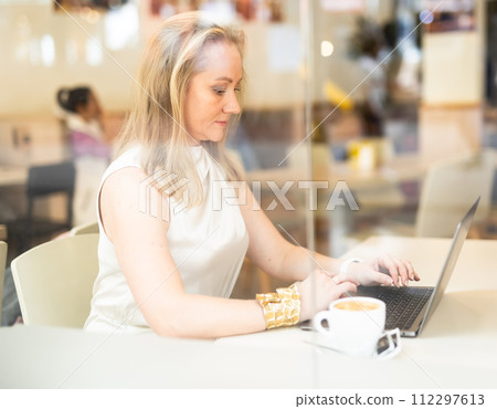 Business woman drinks coffee and works on laptop while sitting at table in cafe 112297613