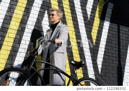 Stylish male in checked coat and sunglasses goes with his retro bicycle by painted urban wall in sunny day. Neutral carbon footprint transportation. Green eco friendly mobility sustainable transport Stylish male in checked coat and sunglasses goes with his retro bicycle by painted urban wall in sunny day. Neutral carbon footprint transportation. Green eco friendly mobility sustainable transport 112298332