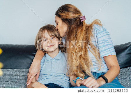 Portrait of happy smiling family at home. Son and mother sitting on the sofa, hugging each other, mom kissing son during home family weekend. Selective focus. 112298333