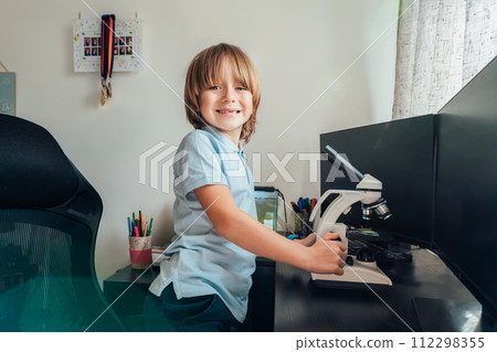 Portrait of inspired Caucasian boy using a microscope at home at his study place. Child curiosity, thirst for knowledge, home learning experience, home remote education concepts. Selective focus. 112298355