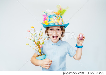 Easter eggs hunt tradition. Emotionally shouting boy kid in Easter hat holds easter tree in pot and shaking chocolate egg in other hand on the white background. Easter decoration and traditions, Easter eggs hunt tradition. Emotionally shouting boy kid in Easter hat holds easter tree in pot and shaking chocolate egg in other hand on the white background. Easter decoration and traditions, 112298358