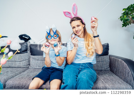 Portraits of happy smiling Caucasian mother and her son in bunny ears having fun with easter eggs at home on Easter day. Family sitting on sofa at home. Family Easter traditions. Selective focus. 112298359