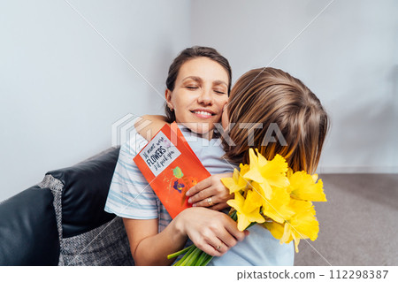 Happy mother's day. Child boy hugging his mother after he congratulates mom and gives her flowers bouquet and handmade postcard. Family holiday and togetherness. Soft selective focus. 112298387