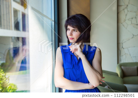 A bright brunette in blue clothes poses by a huge window 112298501