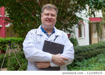 An adult male doctor stands in the shade of a tree with documents in his hands An adult male doctor stands in the shade of a tree with documents in his hands 112298525