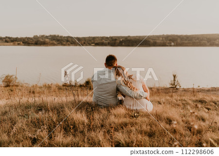 husband with pregnant wife with big belly tummy is resting on bank of river sitting on grass 112298606