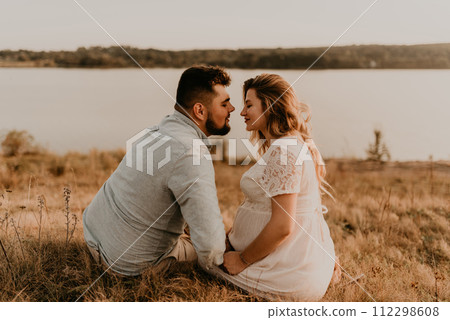 husband with pregnant wife with big belly tummy is resting on bank of river sitting on grass husband with pregnant wife with big belly tummy is resting on bank of river sitting on grass 112298608