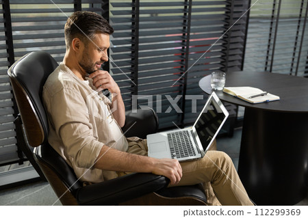 Young bearded man in casual clothes working on laptop Young bearded man in casual clothes working on laptop 112299369
