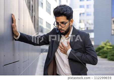 A professional man in a business suit pauses for a moment outside, resting against a wall with a hand over his heart, suggesting contemplation, sincerity, or a health concept. A professional man in a business suit pauses for a moment outside, resting against a wall with a hand over his heart, suggesting contemplation, sincerity, or a health concept. 112299659