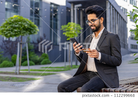 An Indian businessman is engaged with his smartphone while holding a credit card, sitting outside a modern building. This image portrays concepts of mobile banking, online shopping, and digital 112299668