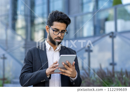 A professional Indian businessman, wearing glasses, is intently using a tablet outside modern glass buildings. The concept of mobile business communication in a global setting is depicted. A professional Indian businessman, wearing glasses, is intently using a tablet outside modern glass buildings. The concept of mobile business communication in a global setting is depicted. 112299689