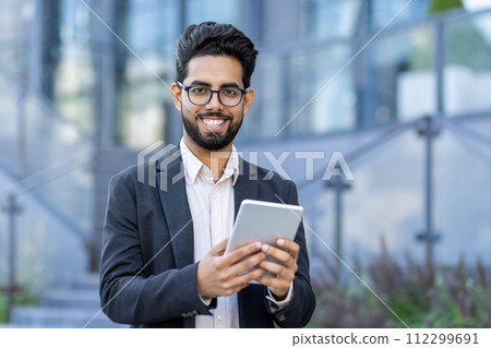 A young businessman in a smart suit engrossed in using his tablet computer. Captured outside a contemporary office setting, symbolizing global business and connectivity. 112299691