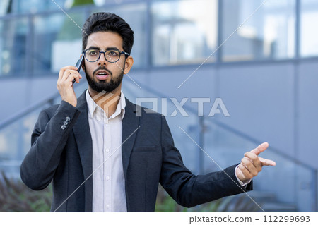 Young, professional businessman in a suit engaging in a conversation on his smartphone outside a modern office building. Young, professional businessman in a suit engaging in a conversation on his smartphone outside a modern office building. 112299693