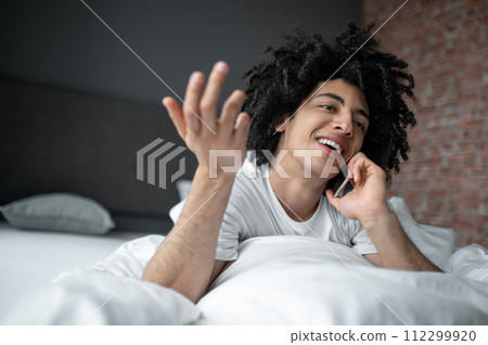 Young man lying in bed and talking on his phone Young man lying in bed and talking on his phone 112299920