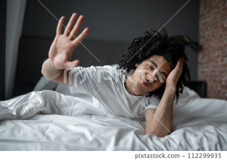 Young curly-haired man lying in bed and looking relaxed and sleepy Young curly-haired man lying in bed and looking relaxed and sleepy 112299935