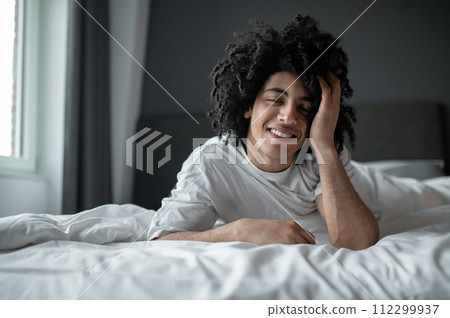Young curly-haired man lying in bed and looking relaxed and sleepy 112299937