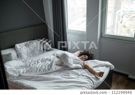Brunette young man lying in bed and stretching after sleep Brunette young man lying in bed and stretching after sleep 112299948