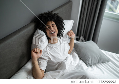 Curly-haired brunette young man waking up in a hotel bedroom Curly-haired brunette young man waking up in a hotel bedroom 112299998