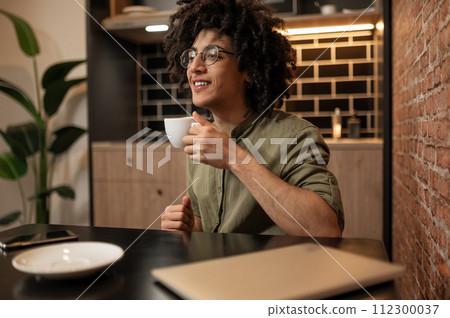 Curly-haired young man sitting at the table and having coffee Curly-haired young man sitting at the table and having coffee 112300037