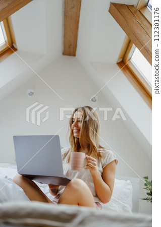 Young woman is sitting on a bed with a laptop and a cup of coffee Young woman is sitting on a bed with a laptop and a cup of coffee 112301176
