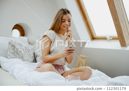 Young smiling woman making notes in note pad while sitting on bed at home 112301179