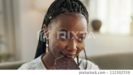 Home, eating and black woman on sofa, chocolate bar and dessert with happiness, relax and sugar snack in a lounge. African person, apartment or girl with candy, relaxing and dessert with comfort food 112302559