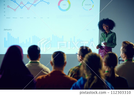 Young African-American woman, speaker at industry event presenting data analytics to group of attentive professionals in conference room. 112303310