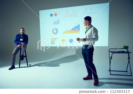 Two men present at business meeting, one seated listening, other standing, gesturing to data chart in conference room. Two men present at business meeting, one seated listening, other standing, gesturing to data chart in conference room. 112303317